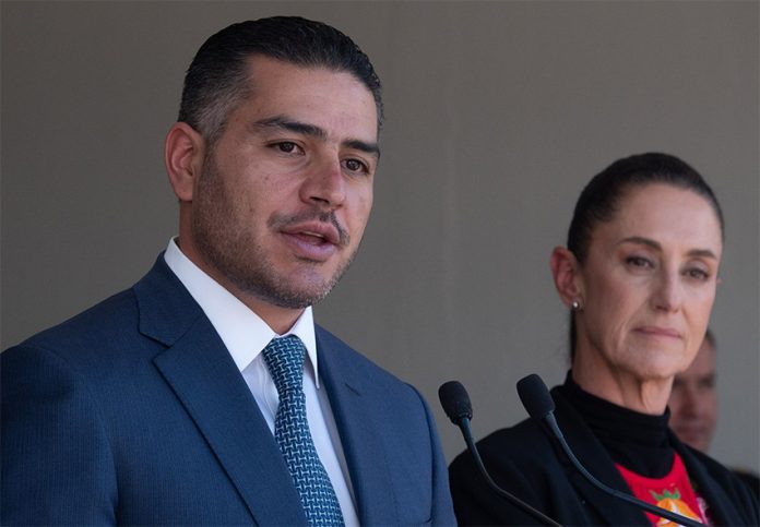 Security Minister García Harfuch speaks at a microphone while Presiden Sheinbaum stands nearby