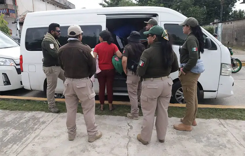 People holding bags of belongings get into a white van, while INM agents in green and khaki uniforms stand behind them watching.