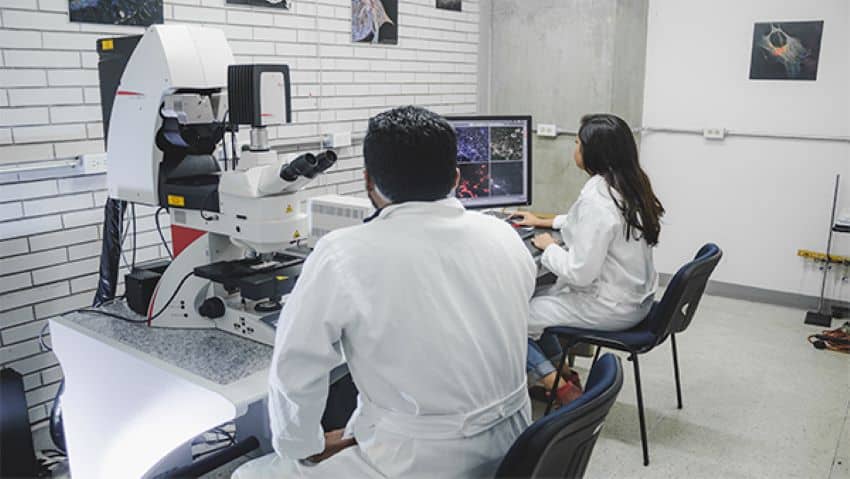 A laboratory room with two researchers in white lab coats, one a man and one a woman. The woman is seated at a desk with a computer and the man is sitting at a table with an advanced microscope.