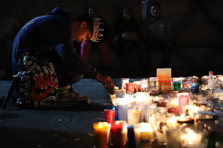 A young man wearing a crown bends down to light one of many candles, whose light illuminates his face