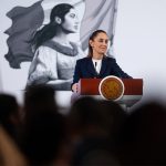 President Claudia Sheinbaum stands at a podium next to a Mexican flag at her morning press briefing