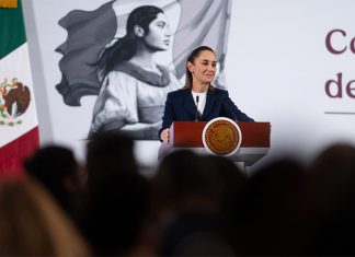 President Claudia Sheinbaum stands at a podium next to a Mexican flag at her morning press briefing