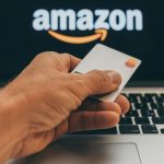 A hand holding a blank Master Card credit card in front of a laptop screen with the Amazon logo in white and orange.