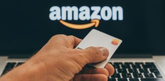A hand holding a blank Master Card credit card in front of a laptop screen with the Amazon logo in white and orange.