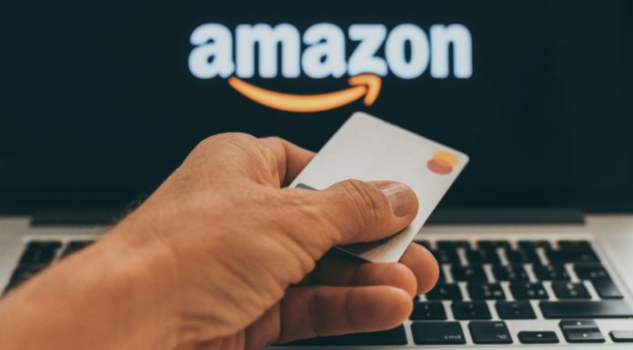 A hand holding a blank Master Card credit card in front of a laptop screen with the Amazon logo in white and orange.