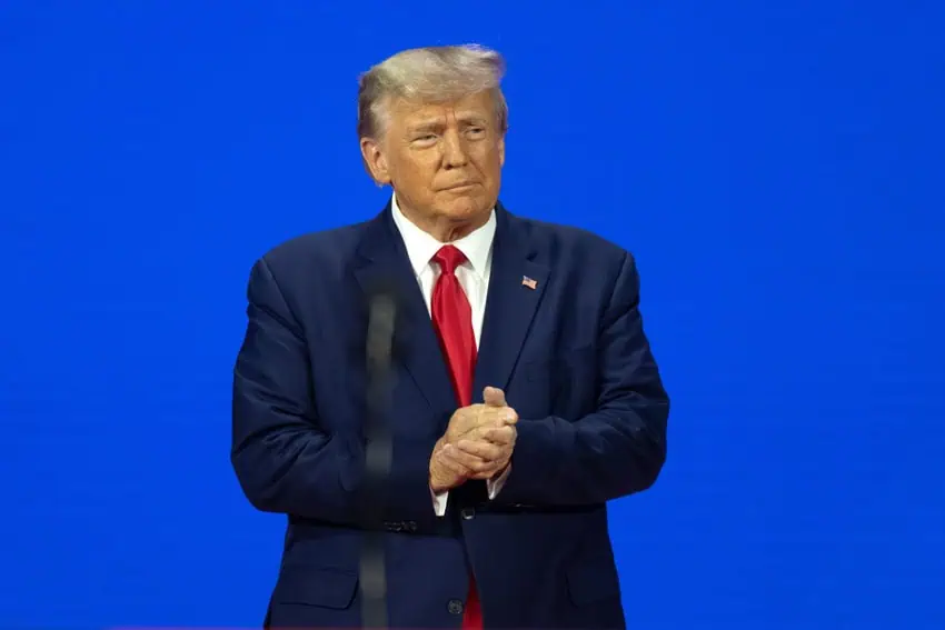 Donald Trump standing against a blue background in a dark blue suit and tie and clapping