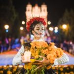 Mexican woman dressed as a Catrina and in white skeletal makeup holding Mexican marigolds