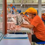 Female employees of a textile factory in Mexico. The women are at work, sitting in rows at tables with industrial sewing machines. They are wearing orange t-shirts and matching orange hats under which their hair is tucked.