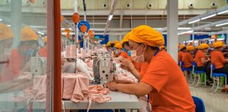 Female employees of a textile factory in Mexico. The women are at work, sitting in rows at tables with industrial sewing machines. They are wearing orange t-shirts and matching orange hats under which their hair is tucked.