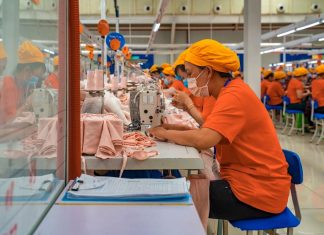 Female employees of a textile factory in Mexico. The women are at work, sitting in rows at tables with industrial sewing machines. They are wearing orange t-shirts and matching orange hats under which their hair is tucked.