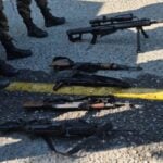 High caliber automatic rifles laid in a row on a street in Mexico. In view nearby are the legs of people in soldier's boots and military fatigues.