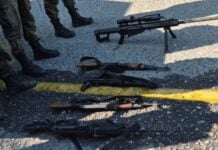 High caliber automatic rifles laid in a row on a street in Mexico. In view nearby are the legs of people in soldier's boots and military fatigues.