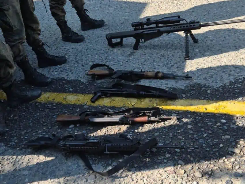 High caliber automatic rifles laid in a row on a street in Mexico. In view nearby are the legs of people in soldier's boots and military fatigues.