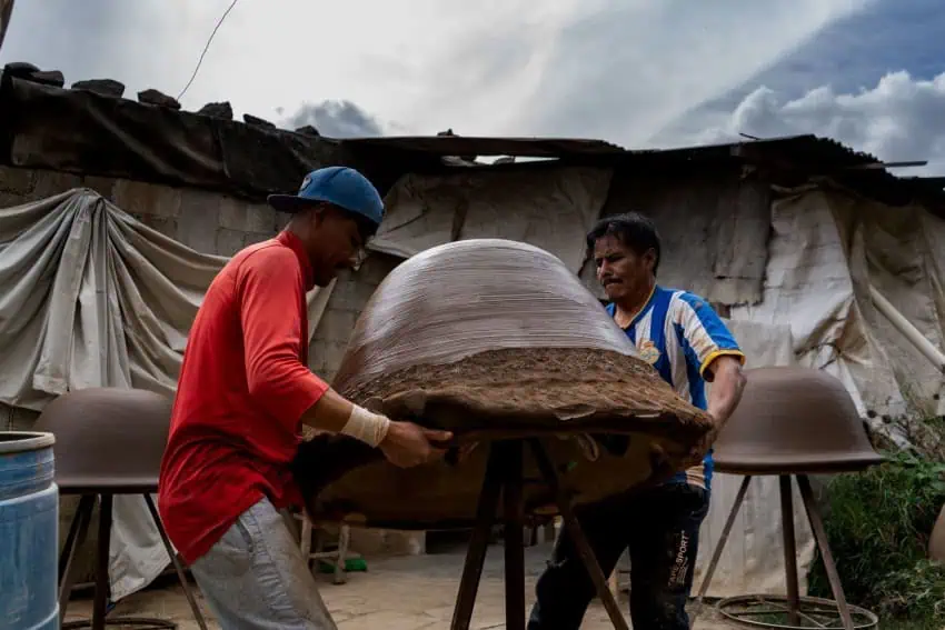 Two Mexican workers lifting a large, heavy pottery project in the shape of a bowl or a bell on an outdoor site in Tlaxcala