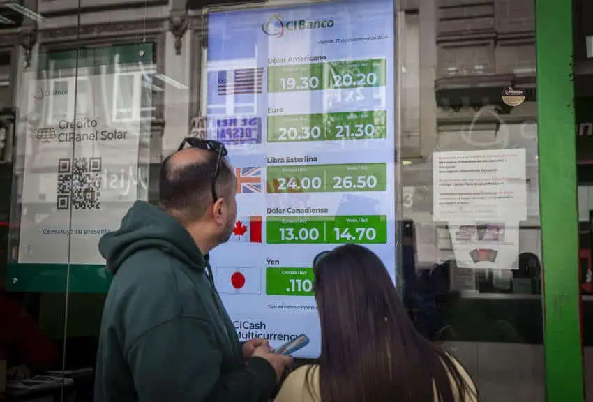 A man and a woman looking at a sign in a bank window listing the rate of the Mexican peso against the US dollar, the euro, the British pound sterling, the Canadian dollar and the Japanese yen.