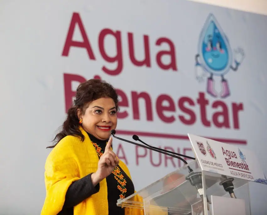 Mexico City Mayor Clara Brugada standing behind a plexiglass podium at a press conference. Behind her is a giant sign that says "Agua Bienestar"