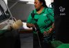 Mexican woman wearing Pemex uniform holding a gas station pump as she prepares to put it into a car's gas tank.