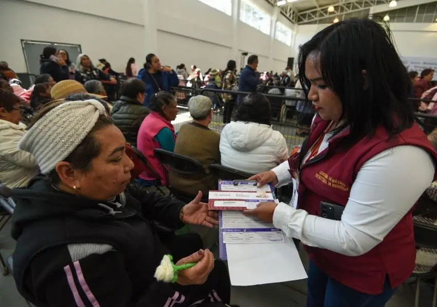 Older Mexican woman receiving paperwork from a Mexican government employee in a crowded event hall.