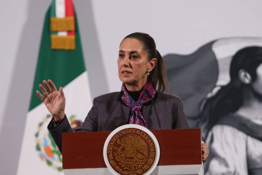 Claudia Sheinbaum at a press conference, standing at the presidential podium, with her right hand raised, palm facing the camera.