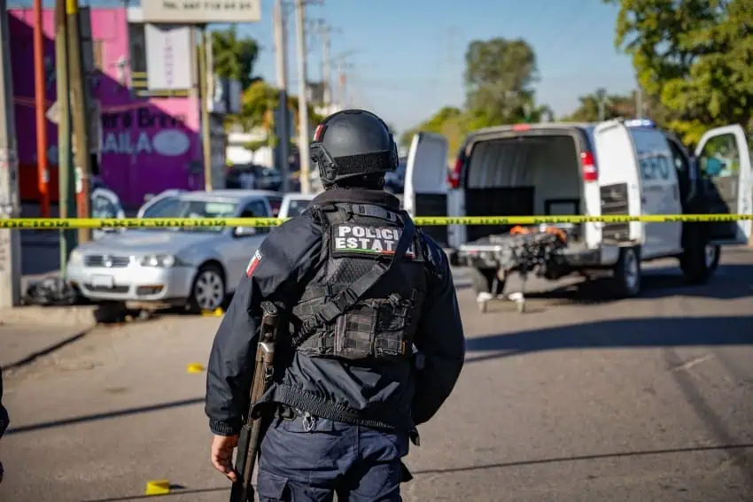 Sinaloa state police officer in black militarized uniform guarding a crime scene on a street in Culiacan, Sinaloa, that is cordoned off with yellow caution tape. In the background is a forensic van with and yellow evidence marker cones on the street.