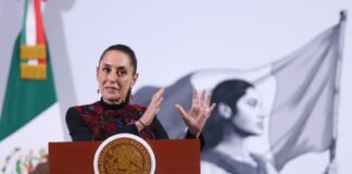Claudia Sheinbaum at a press conference, standing behind the presidential podium, gesturing with both hands parallel in front of her, as if to show the size of something.