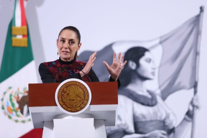 Claudia Sheinbaum at a press conference, standing behind the presidential podium, gesturing with both hands parallel in front of her, as if to show the size of something.