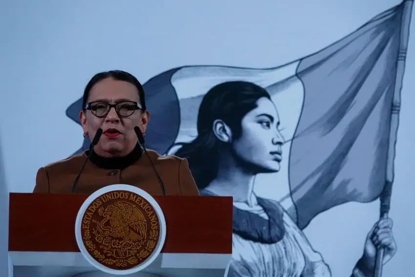 Rosa Icela Rodriguez standing at the presidential podium speaking to reporters. She is wearing a brown buttoned-up wool blazer and black framed glasses.