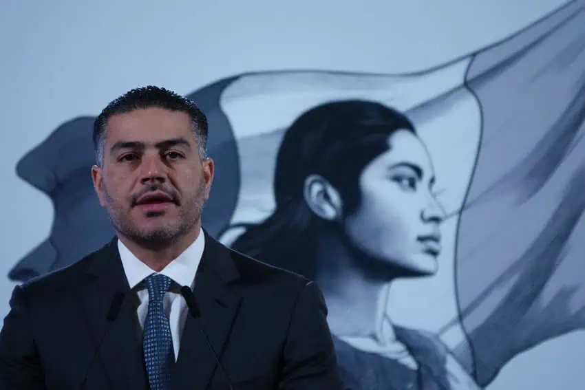 Mexican Security Minister Omar Garcia Harfuch onstage at presidential press conference. Behind him is the Sheinbaum administration's mascot, an illustration of a young Indigenous Mexican woman in front of Mexico's flag.