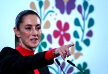 President Claudia Sheinbaum at a press conference in the National Palace, wearing a red turtleneck and a black blazer and pointing her index finger out at a spot in front of her beyond the camera.