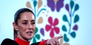 President Claudia Sheinbaum at a press conference in the National Palace, wearing a red turtleneck and a black blazer and pointing her index finger out at a spot in front of her beyond the camera.