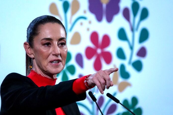 President Claudia Sheinbaum at a press conference in the National Palace, wearing a red turtleneck and a black blazer and pointing her index finger out at a spot in front of her beyond the camera.