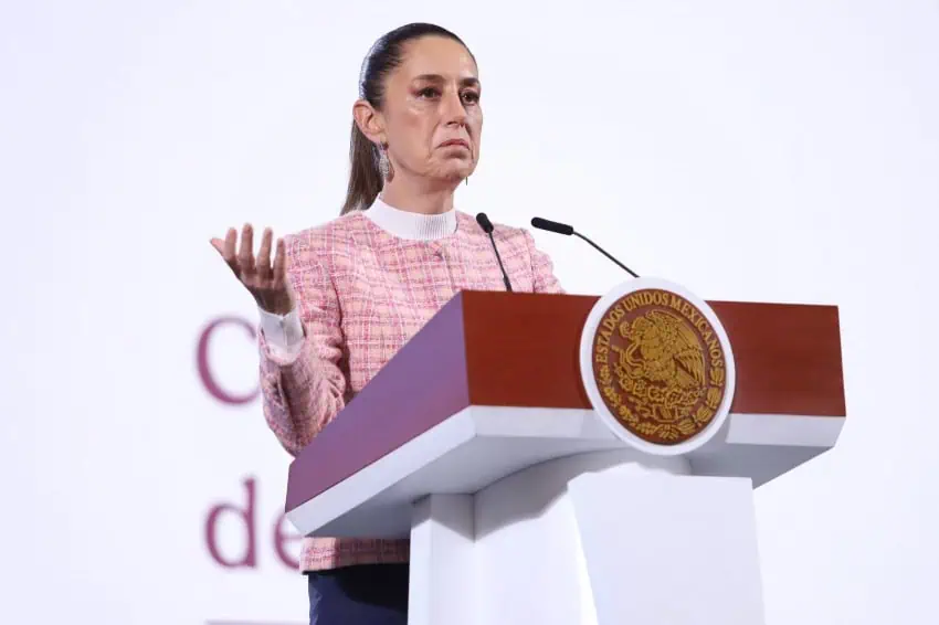 President Claudia Sheinbaum making a shrugging gesture with her right hand at the presidential podium while speaking to reporters during a press conference. She is wearing a pink tartan blazer and a white turtleneck.