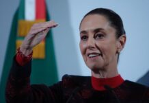 President Claudia Sheinbaum at a press conference. She has her right hand raised to eye level as she makes a point to reporters.