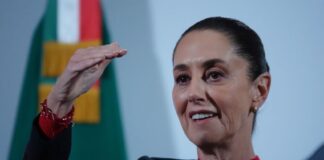 President Claudia Sheinbaum at a press conference. She has her right hand raised to eye level as she makes a point to reporters.
