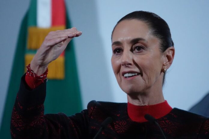 President Claudia Sheinbaum at a press conference. She has her right hand raised to eye level as she makes a point to reporters.