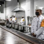 Mexican soldier in a military chef's uniform standing in an industrial kitchen with his hands folded in front of him. In the room are several others dressed just like him, waiting for people to arrive.