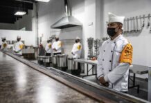 Mexican soldier in a military chef's uniform standing in an industrial kitchen with his hands folded in front of him. In the room are several others dressed just like him, waiting for people to arrive.