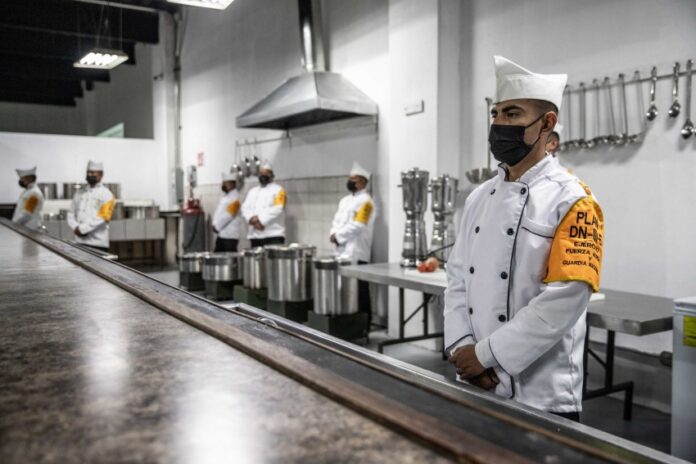 Mexican soldier in a military chef's uniform standing in an industrial kitchen with his hands folded in front of him. In the room are several others dressed just like him, waiting for people to arrive.