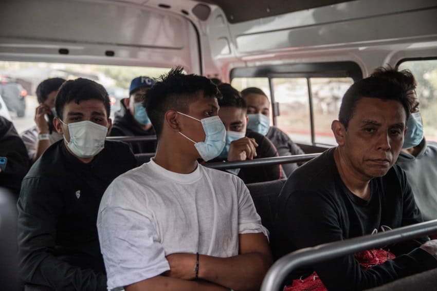 Group of male migrants sitting on a bus, many with COVID-style masks on their faces.