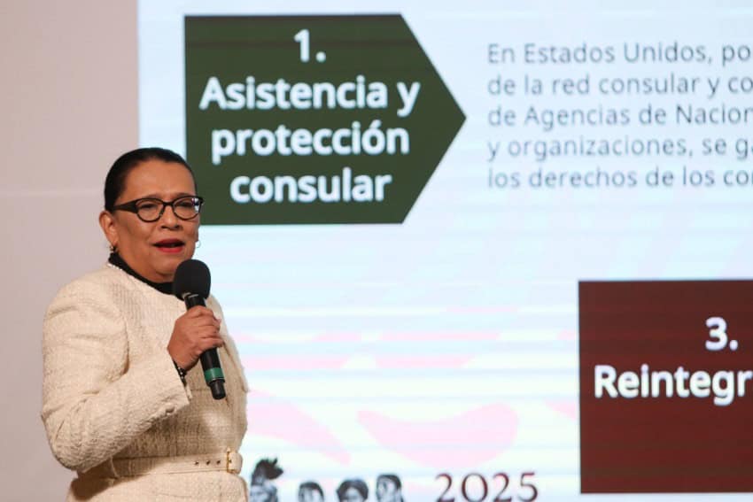 Mexico's Interior Minister Rosa Icela Rodriguez standing in front of a projection screen with a microphone in her right hand as she speaks to reporters. She's wearing a white sweater dress .