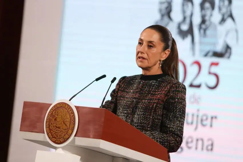 President Claudia Sheinbaum standing at the presidential podium during a press conference, in a sweater of various dark colors and looking out at reporters.