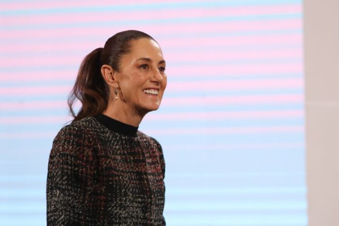 President Claudia Sheinbaum in a dark multicolored sweater smiling at reporters during a press conference at the National Palace press briefing room