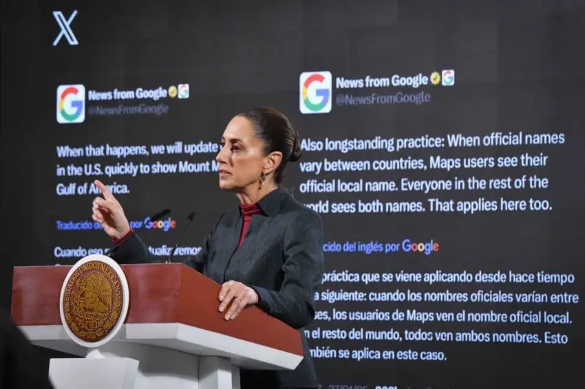 Mexican President Claudia Sheinbaum in a dark suit standing at the presidential podium during a press conference in the National Palace press briefing room. She is standing in front of a projection screen displaying posts from Google's X social media page about the Gulf of Mexico's impending name change on Google Maps