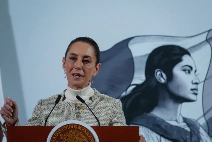 Claudia Sheinbaum standing at the presidential podium in Mexico's National Palace during a press briefing. She's in the middle of speaking to reporters with one closed hand resting on the podium. She's wearing a beige and white blazer and a white turtleneck and her hair is in a ponytail