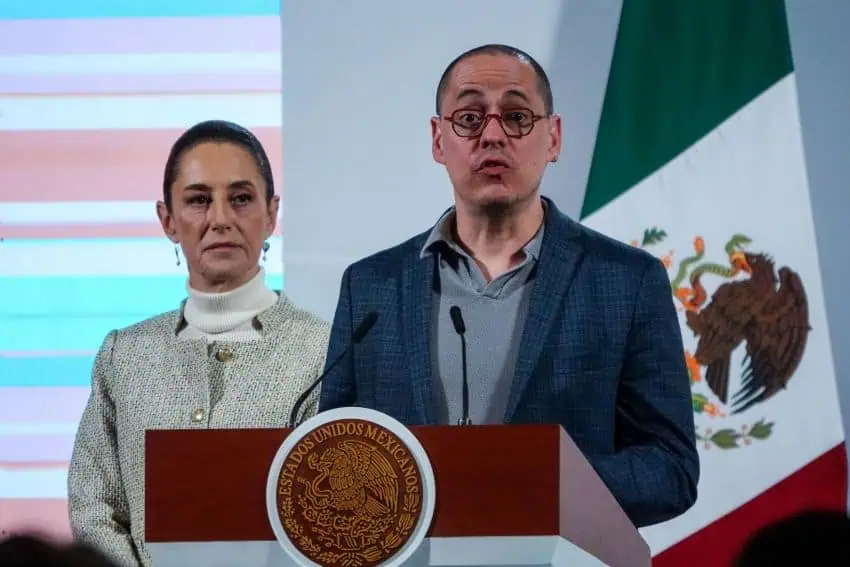 Head of Mexico's Digital Transformation Agency standing at the presidential podium of Mexico, with President Claudia Sheinbaum standing nearby behind him, at a press conferencr at the National Palace. Merino's mouth is puckered and his eyes are wide. Sheinbaum is unsmiling as she looks out at the audience while Merino speaks