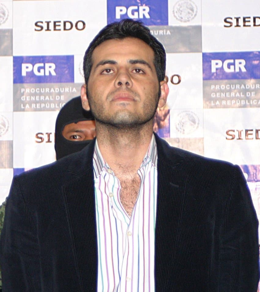 Vicente Zambada Niebla, son of Sinaloa Cartel cofounder Ismael Zambada. He is staring upward at the camera in front of a wall covered with logos of Mexican federal police and the attorney general's office. He is wearing a vertical striped Oxford shirt under a dark blue blazer while a federal police officer with his face covered by a black balaclava looks on from behind him.