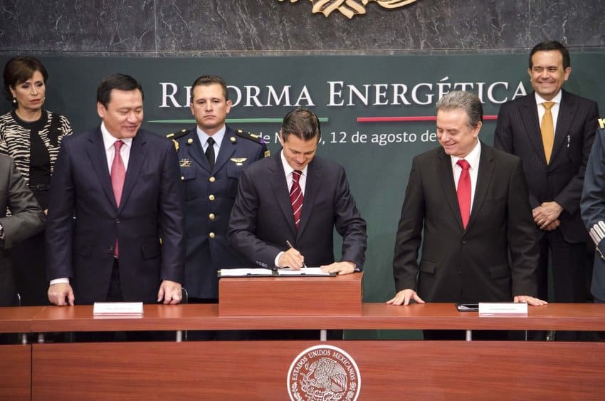 Mexico's former president Enrique Pena Nieto signing a document at a table with the official seal of Mexico in front. He is surrounded by other Mexican politicians and a member of the military. Behind him is a sign saying Reforma Energetica.