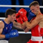 Two men boxing in a white boxing ring. One is wearing red gloves and the other blue. Both gloves have the Paris Olympics logo on them. The boxer in blue is Marco Verde of Mexico and the one in red is Lewis Richardson of the U.K.