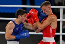 Two men boxing in a white boxing ring. One is wearing red gloves and the other blue. Both gloves have the Paris Olympics logo on them. The boxer in blue is Marco Verde of Mexico and the one in red is Lewis Richardson of the U.K.