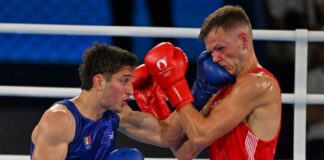 Two men boxing in a white boxing ring. One is wearing red gloves and the other blue. Both gloves have the Paris Olympics logo on them. The boxer in blue is Marco Verde of Mexico and the one in red is Lewis Richardson of the U.K.